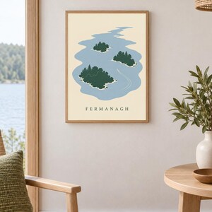 May include: A framed wall art print featuring a stylized illustration of a blue lake with three dark green islands and a small white boat leaving a wake. The text FERMANAGH is printed in black capital letters at the bottom of the cream background.