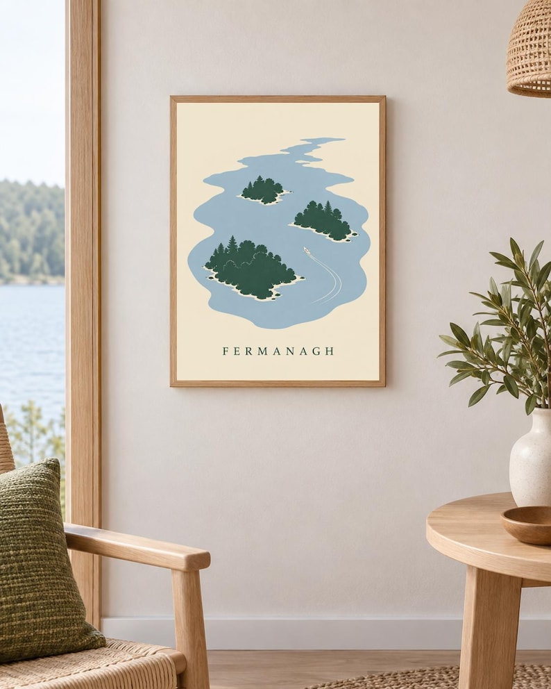 May include: A framed wall art print featuring a stylized illustration of a blue lake with three dark green islands and a small white boat leaving a wake. The text FERMANAGH is printed in black capital letters at the bottom of the cream background.
