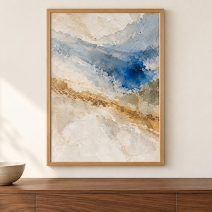 Puede incluir: Una pintura abstracta en acuarela enmarcada que presenta lavados fluidos de azul profundo, oro apagado y tonos beige suaves. La obra de arte est&aacute; encerrada en un marco de madera clara, mostrando texturas en capas y transiciones de color org&aacute;nicas.