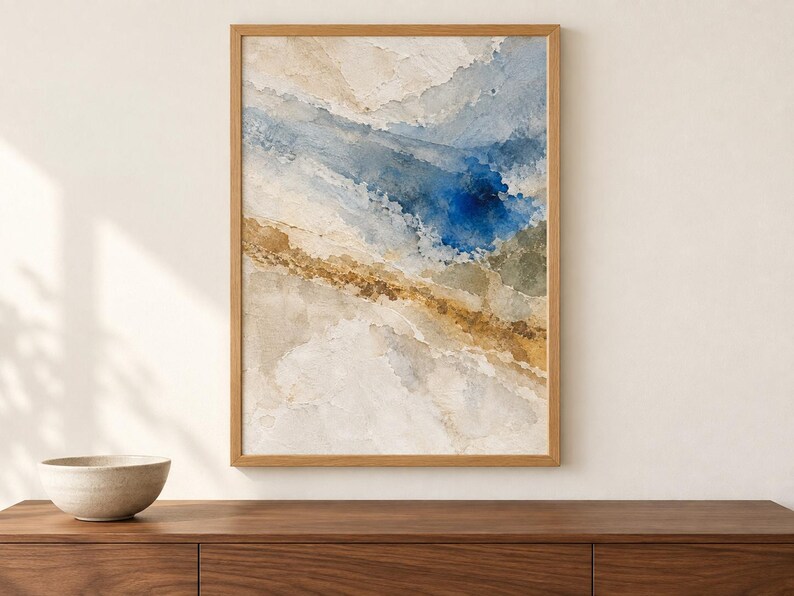 Puede incluir: Una pintura abstracta en acuarela enmarcada que presenta lavados fluidos de azul profundo, oro apagado y tonos beige suaves. La obra de arte est&aacute; encerrada en un marco de madera clara, mostrando texturas en capas y transiciones de color org&aacute;nicas.