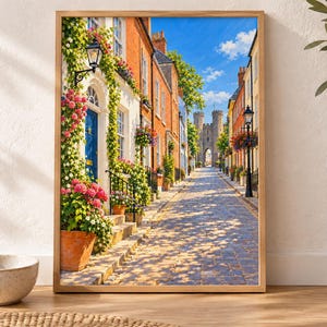May include: A framed wall art print depicting a cobblestone street lined with colorful brick buildings, vibrant flower pots, climbing vines, and a blue front door, leading toward a stone castle gatehouse under a bright blue sky with clouds.