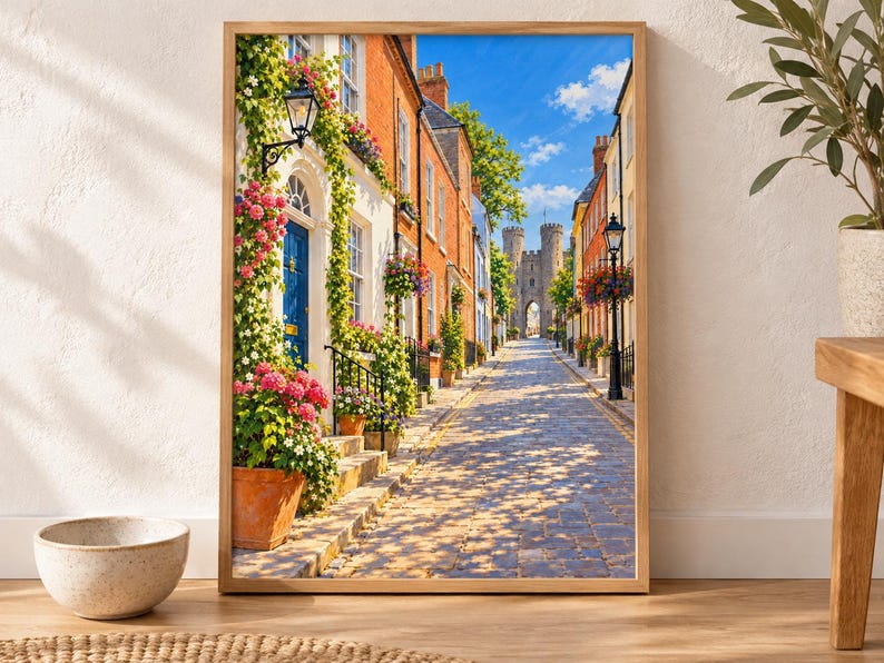 May include: A framed wall art print depicting a cobblestone street lined with colorful brick buildings, vibrant flower pots, climbing vines, and a blue front door, leading toward a stone castle gatehouse under a bright blue sky with clouds.