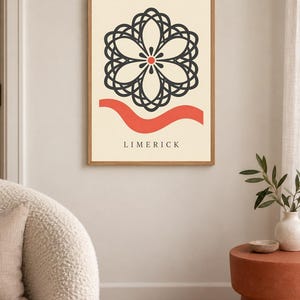 K&ouml;nnte beinhalten: Ein gerahmter Wandkunstdruck mit cremefarbenem Hintergrund, einem zentralen schwarzen geometrischen Blumenmotiv, einem kleinen roten Punkt in der Mitte, einer horizontalen roten Wellenlinie unter dem Motiv und dem Wort LIMERICK in schwarzen Gro&szlig;buchstaben.