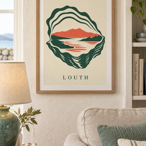 May include: A framed wall art print featuring a stylized dark green oyster shell outline surrounding a landscape illustration of red mountains, a body of water, and a small structure, with the word LOUTH printed in dark green capital letters below.