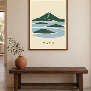 Pu&ograve; includere: Una stampa artistica da parete incorniciata che presenta un'illustrazione paesaggistica stilizzata con una montagna verde scuro, acqua azzurra e piccole isole verdi. Lo sfondo &egrave; color crema e la parola MAYO &egrave; stampata in maiuscolo verde scuro.