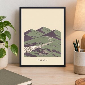 Pu&ograve; includere: Una stampa artistica incorniciata che presenta un'illustrazione paesaggistica stilizzata di colline verdi e cime montuose viola con un muro di pietra che si snoda al centro. La parola DOWN &egrave; stampata in lettere maiuscole nere in basso.