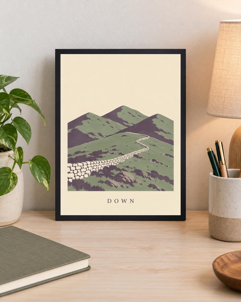 Pu&ograve; includere: Una stampa artistica incorniciata che presenta un'illustrazione paesaggistica stilizzata di colline verdi e cime montuose viola con un muro di pietra che si snoda al centro. La parola DOWN &egrave; stampata in lettere maiuscole nere in basso.