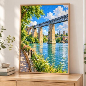 May include: A framed wall art print depicting a stone and metal railway bridge spanning a wide blue river. The scene includes lush green trees, a stone embankment with colorful flowers, and a bright blue sky filled with white clouds.