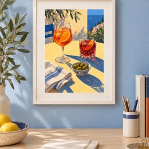 May include: A framed art print depicting a yellow table with an orange cocktail in a stemmed glass, a red cocktail in a tumbler, a small bowl of green olives, and a white napkin with blue stripes, set against a coastal Mediterranean backdrop.