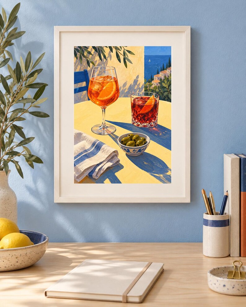 May include: A framed art print depicting a yellow table with an orange cocktail in a stemmed glass, a red cocktail in a tumbler, a small bowl of green olives, and a white napkin with blue stripes, set against a coastal Mediterranean backdrop.