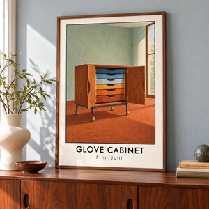 May include: A framed art print titled GLOVE CABINET by Finn Juhl, featuring a wooden cabinet with open doors revealing six colourful drawers in shades of blue, yellow, and orange, set on a dark orange floor against a light green wall.