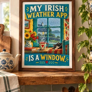 May include: A framed wall art print featuring a rainy window scene with the text MY IRISH WEATHER APP IS A WINDOW. The illustration shows a black and white cat sleeping on a red plaid blanket, a mug with a red heart, and a vase of sunflowers.