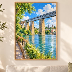 May include: A framed wall art print depicting a stone and metal railway viaduct spanning a wide blue river. The scene includes a sunlit riverside path with green foliage, blooming flowers, and a distant town skyline under a bright blue sky with clouds.