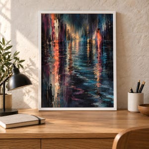 Op de afbeelding: Een ingelijste verticale wandkunstprint met een abstract stadsgezicht bij nacht met levendige oranje, diepblauwe en paarse verticale penseelstreken die reflecteren op een donker wateroppervlak, gepresenteerd in een witte lijst op een houten bureau.