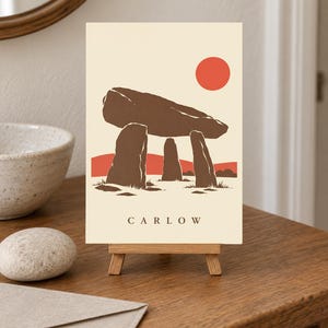 May include: A rectangular print on a small wooden easel featuring a stylized illustration of a brown megalithic dolmen against a cream background. A solid red sun sits in the upper right corner, with red hills below. The word CARLOW is printed in black.