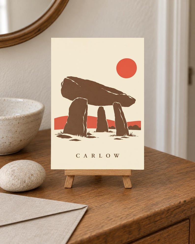 May include: A rectangular print on a small wooden easel featuring a stylized illustration of a brown megalithic dolmen against a cream background. A solid red sun sits in the upper right corner, with red hills below. The word CARLOW is printed in black.
