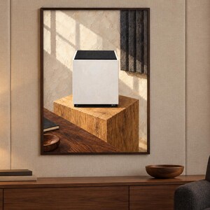 Op de afbeelding: Een witte kubusvormige luidspreker met een zwart bovenpaneel, weergegeven in een ingelijst wandkunstwerk. Het kunstwerk toont de luidspreker rustend op een houten blok, tegen een textuurrijke achtergrond met licht- en schaduwpatronen.