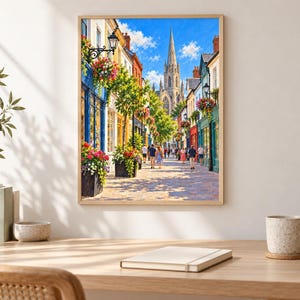 Op de afbeelding: Een ingelijste muurkunstprint met een zonnig Europees straatbeeld met kleurrijke winkelpuien, hangende bloemenmanden en een hoge stenen kerktoren op de achtergrond. Mensen lopen over het geplaveide pad onder een helderblauwe lucht.