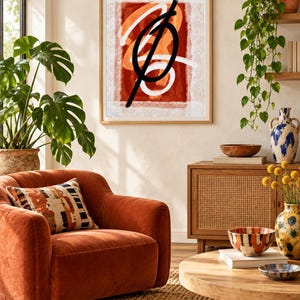 May include: A framed abstract wall art print featuring a bold black linear stroke over layered white and burnt orange shapes. The artwork is displayed in a light wood frame, showcasing a composition of contrasting colors and fluid, gestural lines.