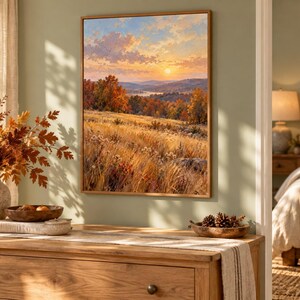 May include: A rectangular wall art print featuring a landscape painting of a golden autumn field at sunset. The scene depicts tall grasses, colorful fall foliage on distant hills, and a soft, glowing sun in a cloudy sky, all framed in a light wood border.