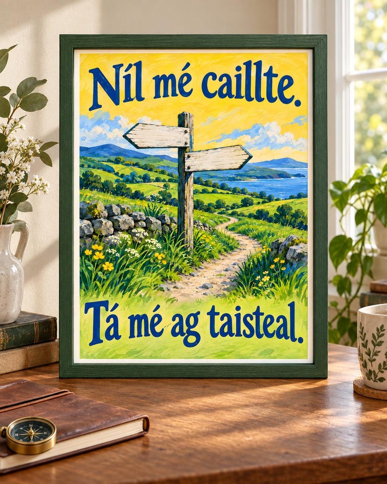 Op de afbeelding: Een ingelijste print met een landschapsschilderij van een zandpad, een houten wegwijzer en groene heuvels onder een gele lucht. De tekst "N&iacute;l m&eacute; caillte." staat bovenaan en "T&aacute; m&eacute; ag taisteal." onderaan in dikke blauwe letters.