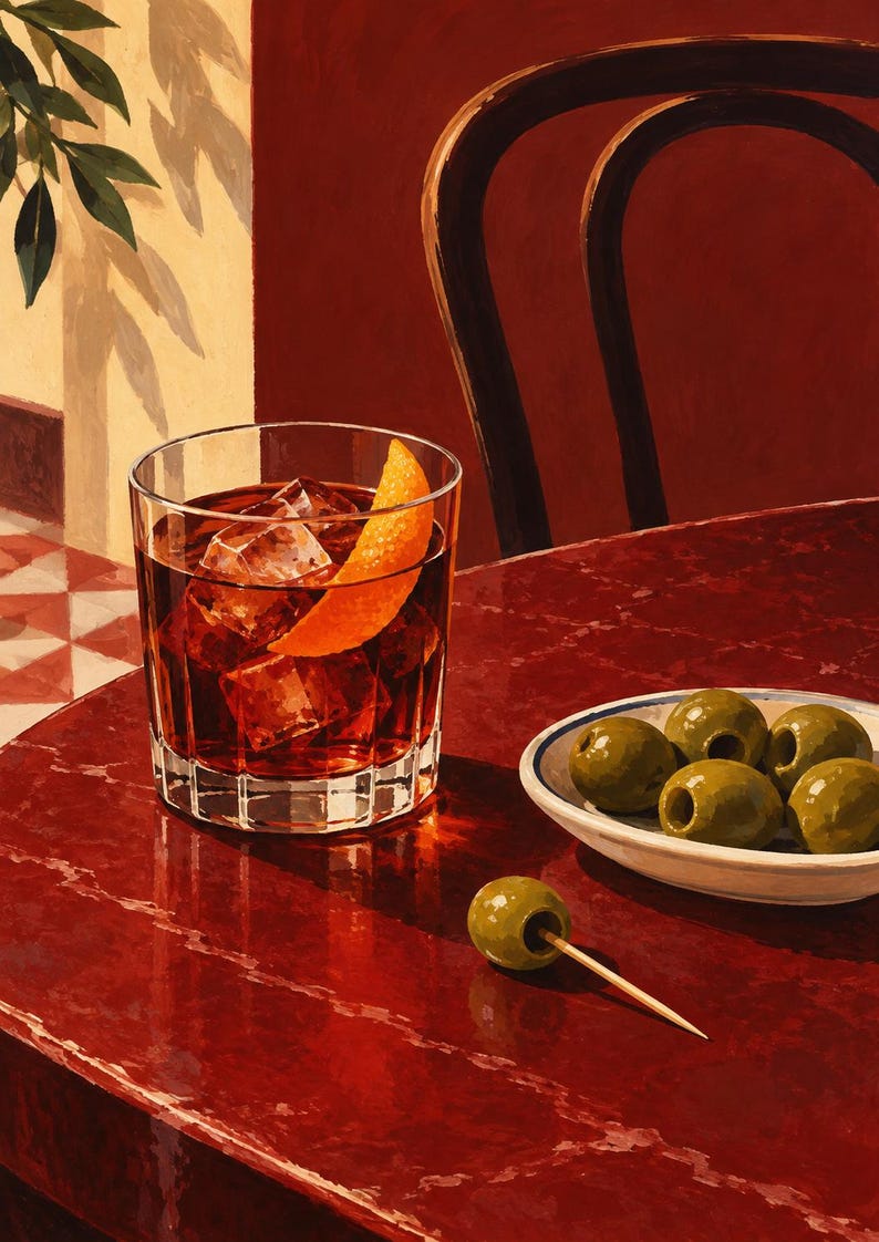 May include: A glass of dark amber liquid with ice cubes and an orange peel garnish sits on a deep red table next to a small white bowl containing five green olives and a single green olive on a wooden toothpick resting on the table surface.