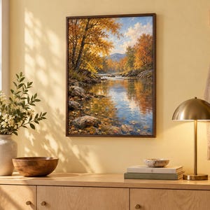 K&ouml;nnte beinhalten: Ein rechteckiger gerahmter Kunstdruck, der eine ruhige Herbstlandschaft mit einem flie&szlig;enden Fluss, felsigen Ufern und leuchtend orangefarbenen und gelben B&auml;umen unter einem blauen Himmel zeigt, montiert an einer hellgelben Wand &uuml;ber einem Schrank.