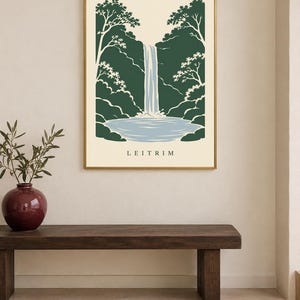K&ouml;nnte beinhalten: Ein rechteckiger Wandkunstdruck in einem goldenen Rahmen mit einer stilisierten Illustration eines Wasserfalls, der in ein Becken st&uuml;rzt, umgeben von dunkelgr&uuml;nen B&auml;umen auf cremefarbenem Grund, mit dem Wort LEITRIM in Schwarz unten.