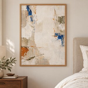 K&ouml;nnte beinhalten: Ein rechteckiges Wandbild in einem hellen Holzrahmen mit einer abstrakten Komposition. Das Kunstwerk zeigt geschichtete Texturen in Off-White-, Creme- und Beiget&ouml;nen, akzentuiert durch kr&auml;ftige Flecken in tiefem Blau und gebranntem Orange.