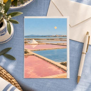 May include: A rectangular greeting card featuring a landscape illustration of salt pans with pink and blue water, a white salt mound, and a distant town under a pale sun. The card is placed on a blue fabric surface next to a white envelope and a pen.