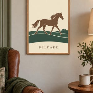 May include: A framed wall art print featuring a brown horse galloping across a dark green landscape against a cream background. The word KILDARE is printed in dark brown capital letters at the bottom of the print, which is encased in a light wood frame.