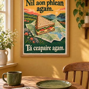 Op de afbeelding: Een ingelijste kunstdruk met een sandwich op een opengevouwen kaart voor een kustlandschap. De tekst luidt bovenaan N&iacute;l aon phlean agam. en onderaan T&aacute; ceapaire agam. De illustratie bevat groene, blauwe en oranje kleurtinten.
