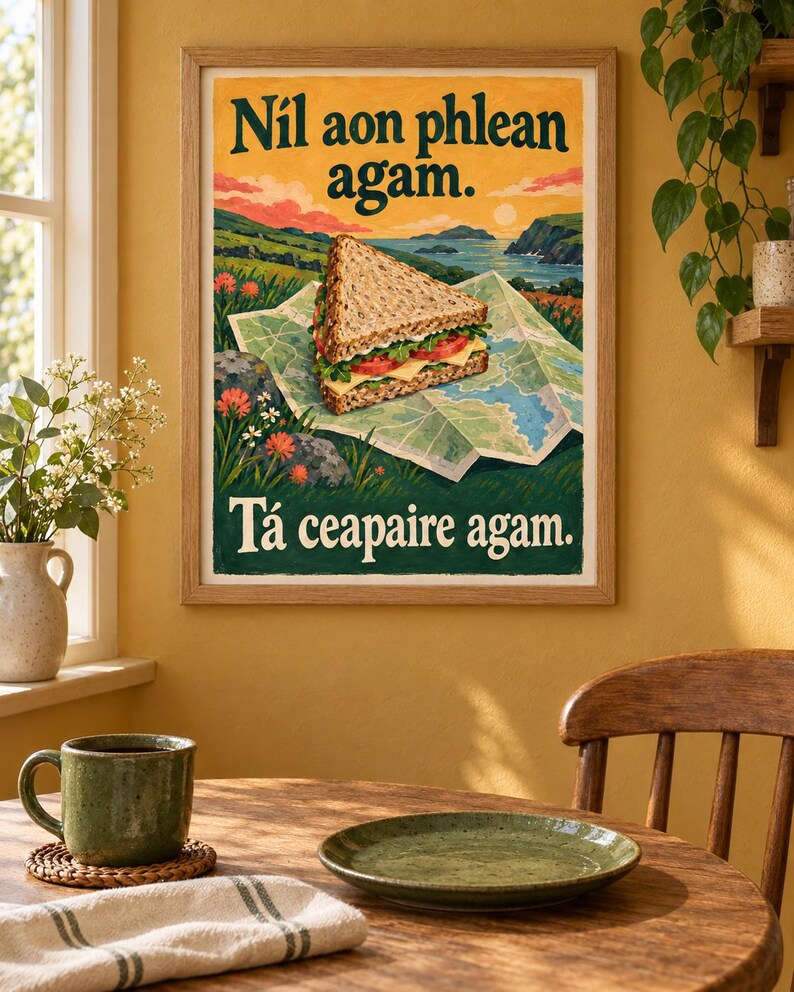 Op de afbeelding: Een ingelijste kunstdruk met een sandwich op een opengevouwen kaart voor een kustlandschap. De tekst luidt bovenaan N&iacute;l aon phlean agam. en onderaan T&aacute; ceapaire agam. De illustratie bevat groene, blauwe en oranje kleurtinten.