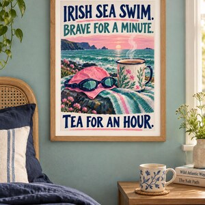 May include: A framed wall art print featuring a coastal scene with a pink swim cap, blue goggles, and a floral mug on a rock. The text reads IRISH SEA SWIM. BRAVE FOR A MINUTE. TEA FOR AN HOUR. The illustration includes a sunset over the ocean.
