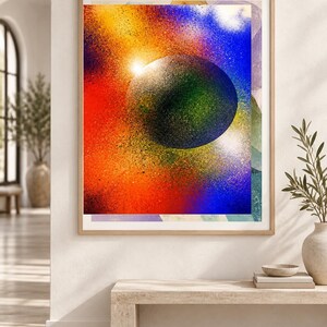 Op de afbeelding: Een ingelijste rechthoekige wandkunstprint met een centrale donkere bol omringd door een levendig, gespoten kleurverloop van rood, oranje, geel en blauw. Het kunstwerk is ingelijst in een licht houten frame met een kosmische uitstraling.
