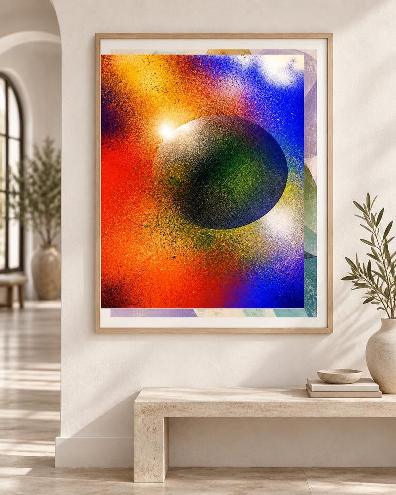 Op de afbeelding: Een ingelijste rechthoekige wandkunstprint met een centrale donkere bol omringd door een levendig, gespoten kleurverloop van rood, oranje, geel en blauw. Het kunstwerk is ingelijst in een licht houten frame met een kosmische uitstraling.