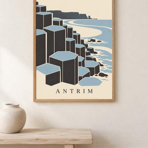 May include: A framed wall art print featuring a stylised illustration of the Giant's Causeway in Antrim. The graphic depicts dark grey hexagonal basalt columns meeting a light blue sea and sandy coastline, with the word ANTRIM printed at the bottom.