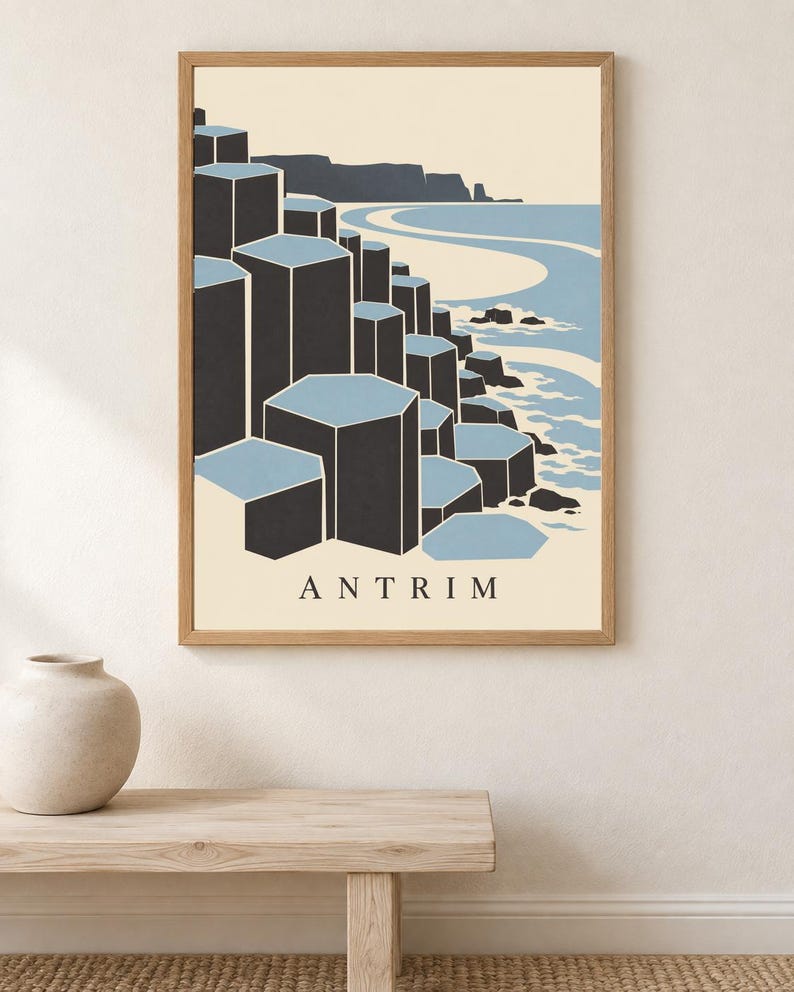 May include: A framed wall art print featuring a stylised illustration of the Giant's Causeway in Antrim. The graphic depicts dark grey hexagonal basalt columns meeting a light blue sea and sandy coastline, with the word ANTRIM printed at the bottom.