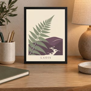 May include: A framed wall art print featuring a stylised green fern leaf in the foreground against a cream background. The lower portion depicts a dark purple mountain landscape with a winding river. The word LAOIS is printed in black at the bottom.