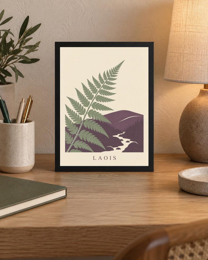 May include: A framed wall art print featuring a stylised green fern leaf in the foreground against a cream background. The lower portion depicts a dark purple mountain landscape with a winding river. The word LAOIS is printed in black at the bottom.
