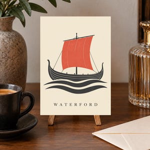 Pode incluir: Uma impress&atilde;o retangular num pequeno cavalete de madeira com um navio viking preto com uma grande vela vermelha, navegando sobre ondas pretas estilizadas. A palavra WATERFORD est&aacute; impressa em letras mai&uacute;sculas pretas no fundo creme.