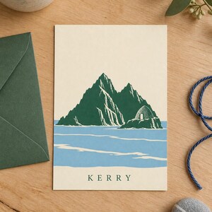 Pu&ograve; includere: Un biglietto d'auguri rettangolare con un'illustrazione serigrafata di montagne verdi e una capanna di pietra a forma di alveare che emerge dall'acqua blu. La parola KERRY &egrave; stampata in lettere maiuscole verde scuro al centro della base del biglietto.