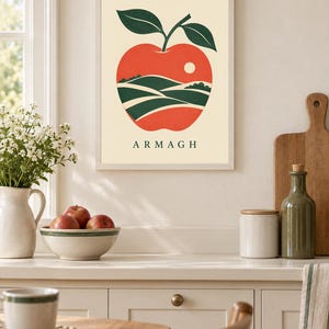 May include: A framed wall art print featuring a stylized red apple with two dark green leaves. Inside the apple shape is a landscape illustration of rolling green hills and a small white sun. The text ARMAGH is printed in black below the apple.