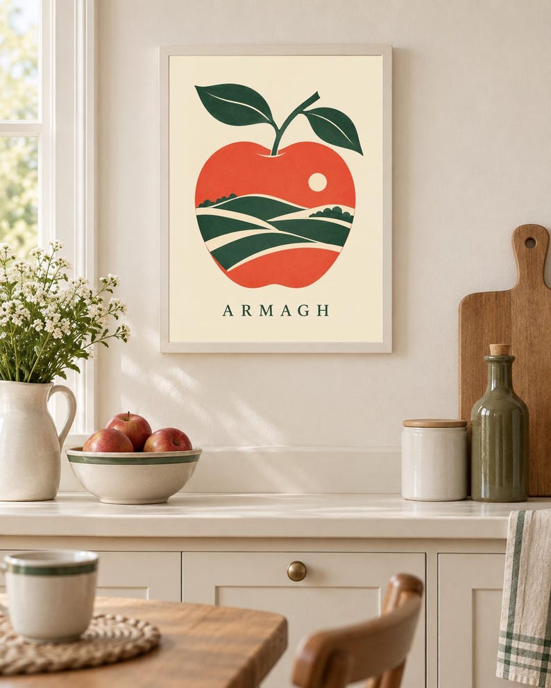 May include: A framed wall art print featuring a stylized red apple with two dark green leaves. Inside the apple shape is a landscape illustration of rolling green hills and a small white sun. The text ARMAGH is printed in black below the apple.