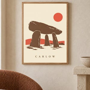May include: A framed wall art print featuring a stylized illustration of a brown megalithic dolmen structure against a cream background. A solid orange sun sits in the upper right corner above a red horizon line. The word CARLOW is printed in black.