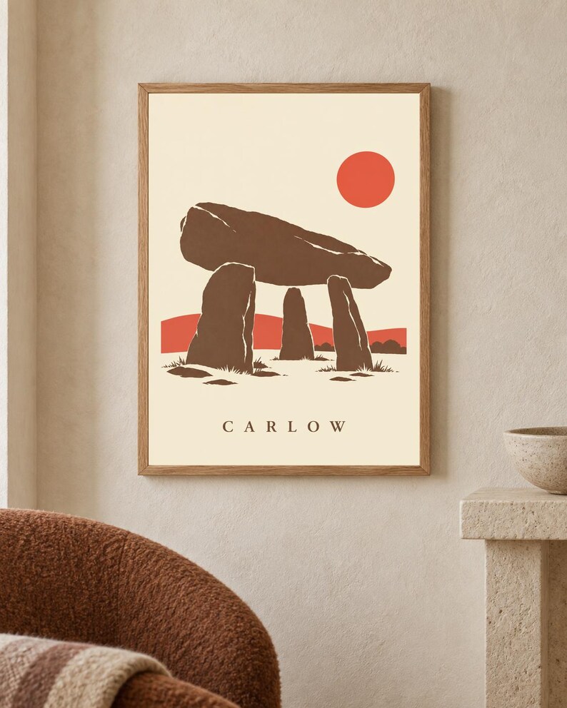 May include: A framed wall art print featuring a stylized illustration of a brown megalithic dolmen structure against a cream background. A solid orange sun sits in the upper right corner above a red horizon line. The word CARLOW is printed in black.
