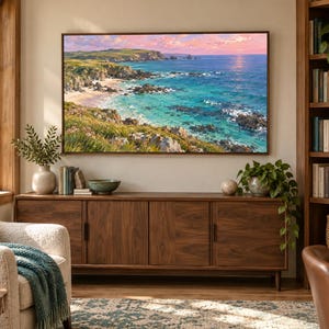 May include: A rectangular wall-mounted television displaying a vibrant coastal landscape painting featuring turquoise ocean waters, rocky cliffs, a sandy beach, and a soft pink sunset sky, framed in a thin dark border and mounted above a wooden sideboard.