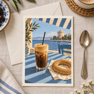 May include: A rectangular art print depicting a glass of iced coffee with a black straw and a sesame seed bagel on a blue table. The scene shows a coastal view with a white tower, blue sea, and striped awning, framed by an olive branch in a vase.