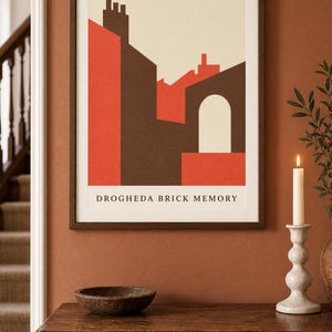 K&ouml;nnte beinhalten: Ein gerahmter Kunstdruck mit einer geometrischen architektonischen Illustration von Geb&auml;uden in Rot-, Braun- und Cremet&ouml;nen. Der Text DROGHEDA BRICK MEMORY ist in Schwarz am unteren Rand des Drucks zu sehen, der in einem dunklen Holzrahmen steckt.