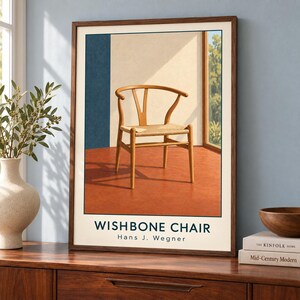 May include: A framed wall art print featuring a light wood Wishbone chair with a woven seat. The illustration depicts the chair in a room with a terracotta floor, a blue wall section, and a window. The text reads WISHBONE CHAIR Hans J. Wegner.